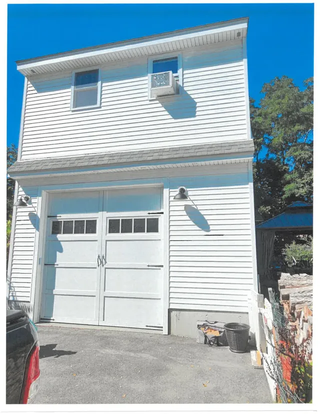 Exterior photo of a white two-story garage or accessory building with double garage doors, a second-story window, and an air-conditioning unit mounted near the upper window. The building is shown from a low angle with a driveway in front and trees visible at the side.