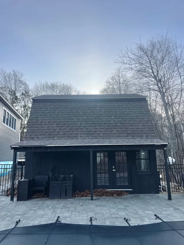 Exterior photograph of a small dark-painted rear accessory structure or shed-like building viewed straight on from the patio, with a shingled gable roof, glass double doors, and surrounding trees and adjacent house visible at the sides.