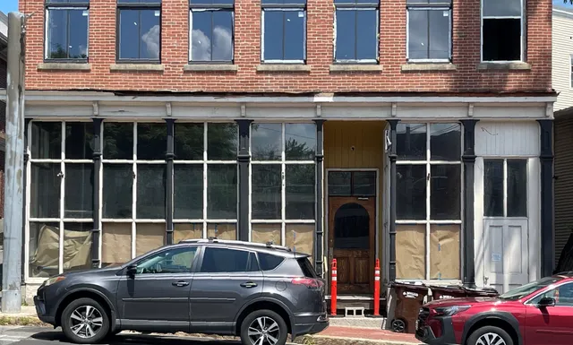 Street-level photo of the existing front facade of 11 Elm St, showing a two-story brick building with large ground-floor storefront windows and a central entrance. Several parked vehicles partially block the lower portion of the building.