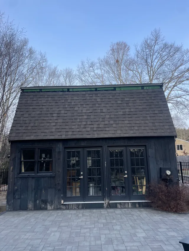Exterior photo of a small dark-clad outbuilding or accessory structure viewed straight on, showing a steep shingled roof, double glass doors, windows, and a paved patio in front with leafless trees behind.