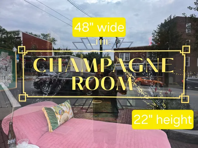Street-level photo looking through a storefront window showing the proposed sign location for "The Champagne Room," with a 48-inch-wide by 22-inch-high sign layout indicated on the glass. The view includes reflections of the street and the interior seating area.