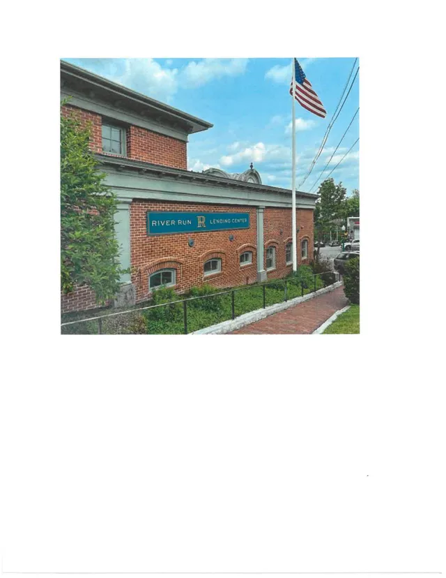 Street-level photo of a brick building with a "River Run Lending Center" sign, seen from an angled view with an American flagpole in the foreground and sidewalk/streetscape along the right side.