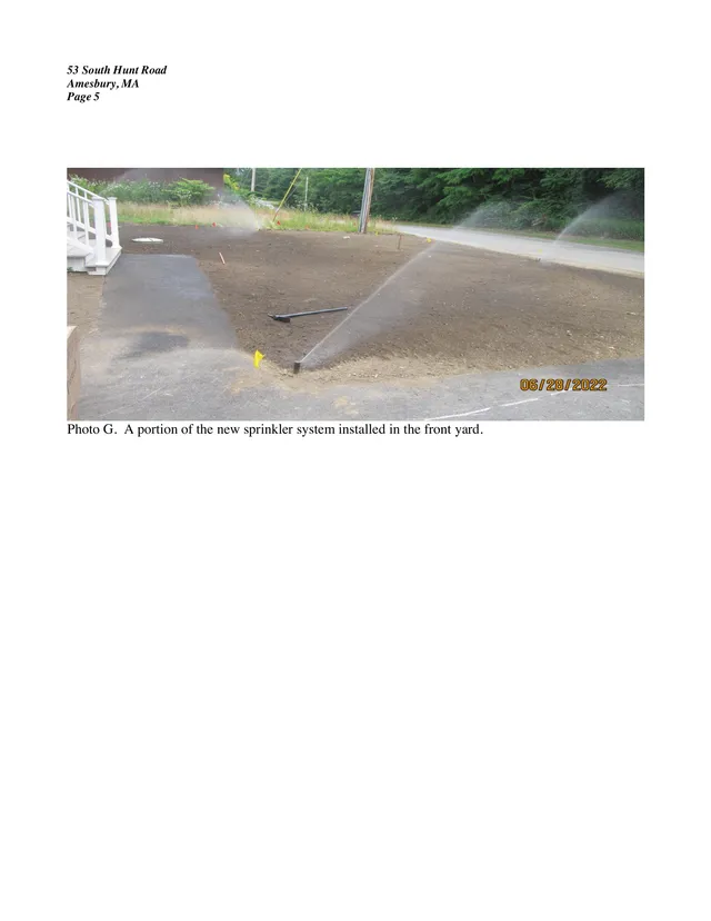 Photograph of the front yard at 53 South Hunt Road showing a partially installed sprinkler system spraying over freshly graded soil near the driveway and road, with trees and utility poles in the background.